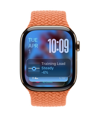 Růžově zlaté Apple Watch s mandarinkovým pleteným řemínkem, v levém horním rohu ciferníku se zobrazuje datum, vpravo nahoře čas psaný velkými číslicemi, dole widget Tréninková zátěž, to všechno v průsvitnémm Liquid Glass designu