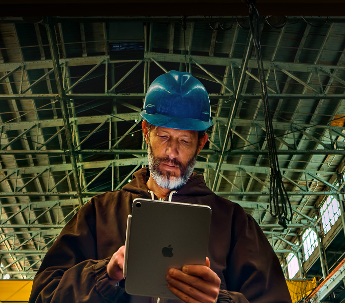 iPad, color argento, con una Apple Pencil agganciata, tenuto in mano da una persona vestita con una giacca marrone e un elmetto blu, al lavoro in un ambiente industriale con travi metalliche sospese