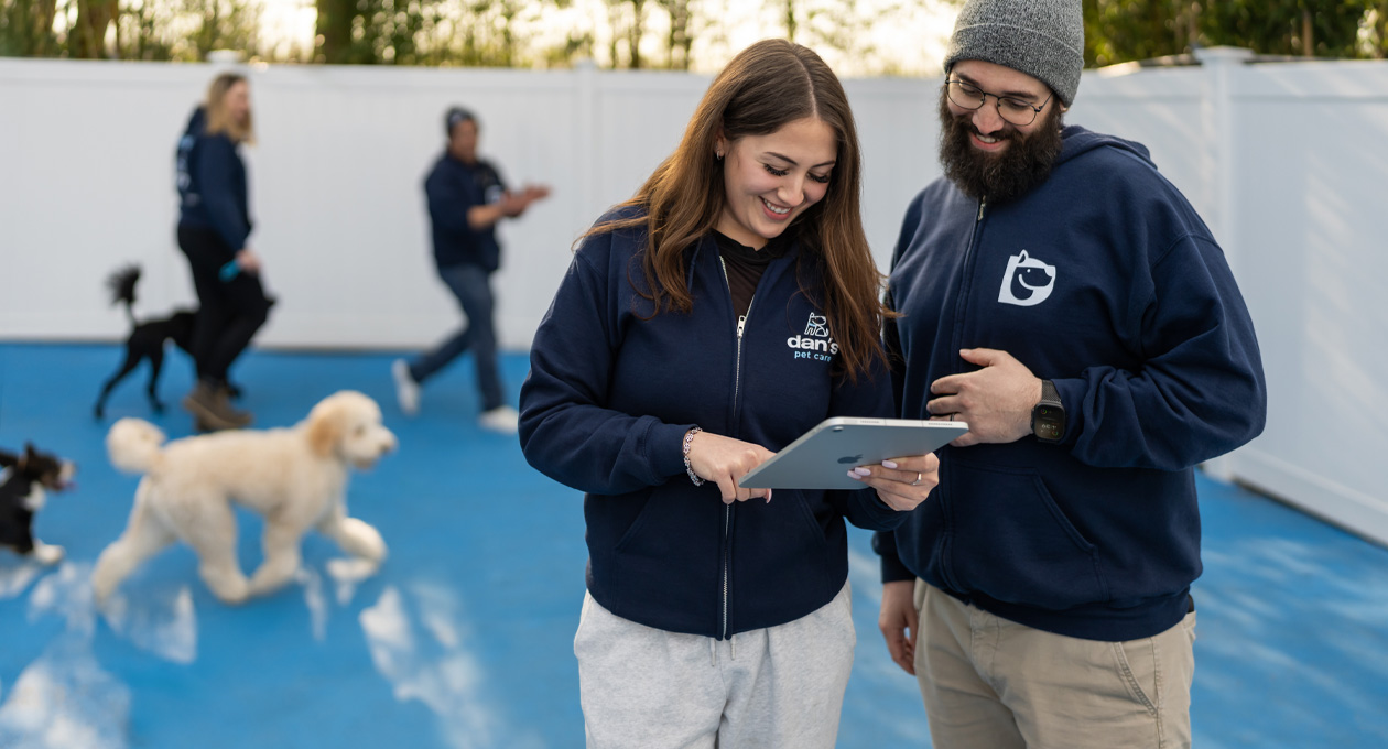 Due dipendenti con felpe griffate abbinate guardano insieme un iPad in un’area cani, sullo sfondo si notano cani e lo staff