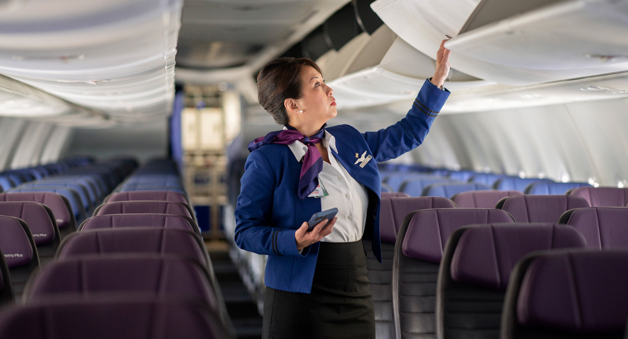 Un’assistente di bordo tiene in mano un iPhone mentre apre una cappelliera in un aereo vuoto