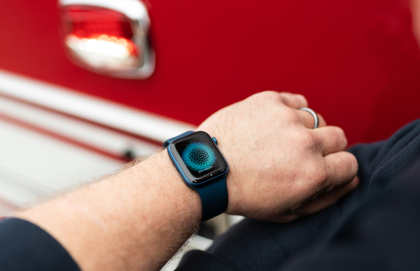 佩戴著 Apple Watch 的個人手臂特寫，錶面顯示英文訊息 Now Inhale 提醒進行吸氣，不遠處有一輛消防車